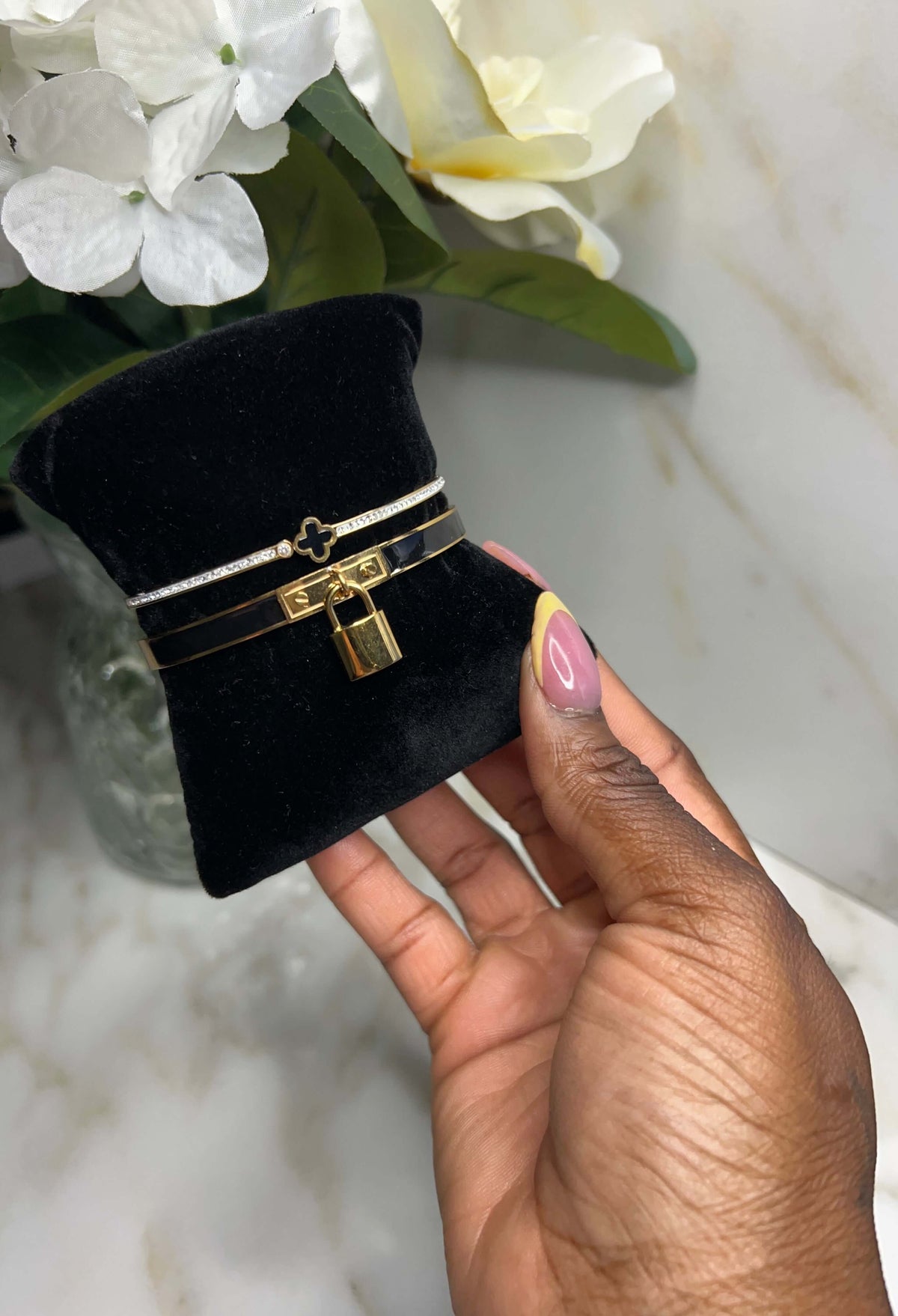 A close-up of a hand holding a black velvet jewelry display cushion with a gold and silver bracelet featuring a padlock motif, set against a marble surface with white flowers in the background.