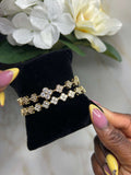 A pair of gold-tone floral bracelets with clear rhinestones displayed on a black velvet jewelry stand, held by hands with pink and yellow manicured nails, with white flowers in the background.