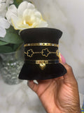 A hand holds a black velvet jewelry display hat showcasing three gold-tone bracelets. The bracelets feature a Roman numeral VIII band, a quatrefoil chain band, and a black band with a heart clasp.