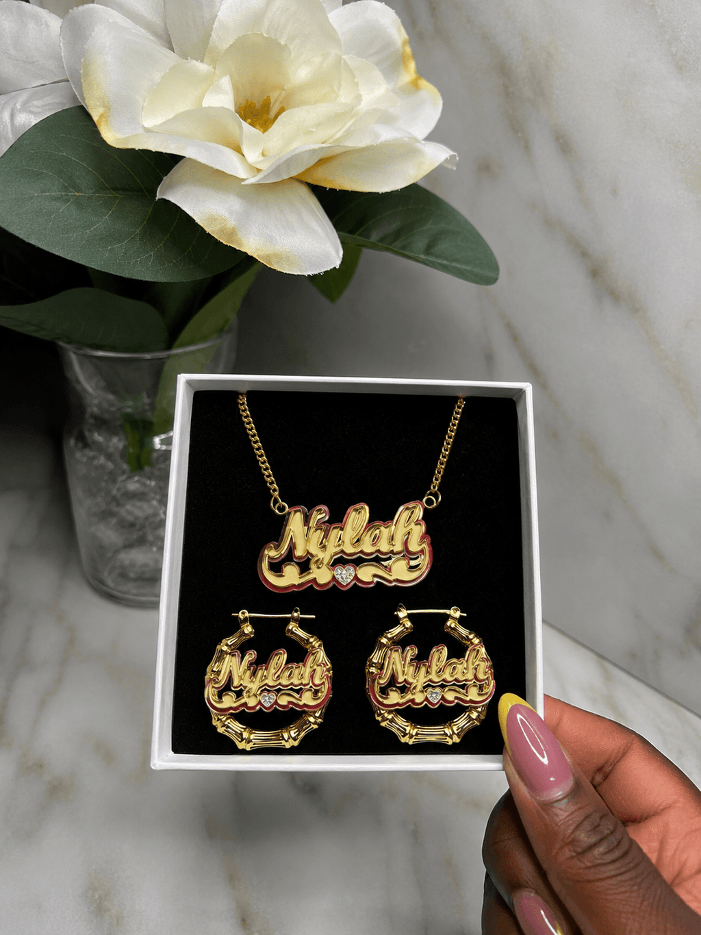 A gold-tone personalized jewelry set with a necklace and matching hoop earrings displayed in a white gift box. The name 'Niklah' is written in cursive across the pendant and earrings, with small clear stones as accents.