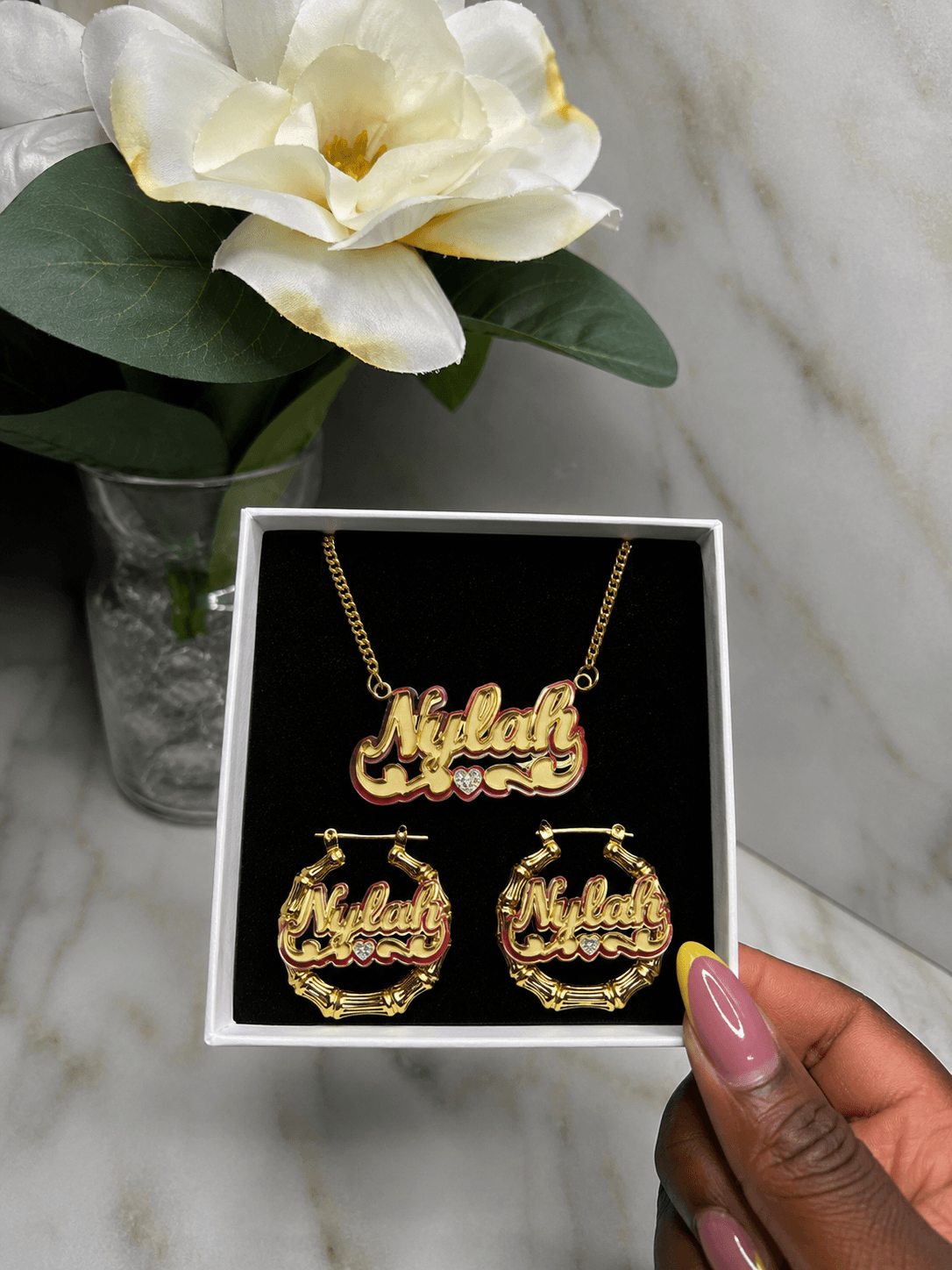 A gold-tone personalized jewelry set with a necklace and matching hoop earrings displayed in a white gift box. The name 'Niklah' is written in cursive across the pendant and earrings, with small clear stones as accents.